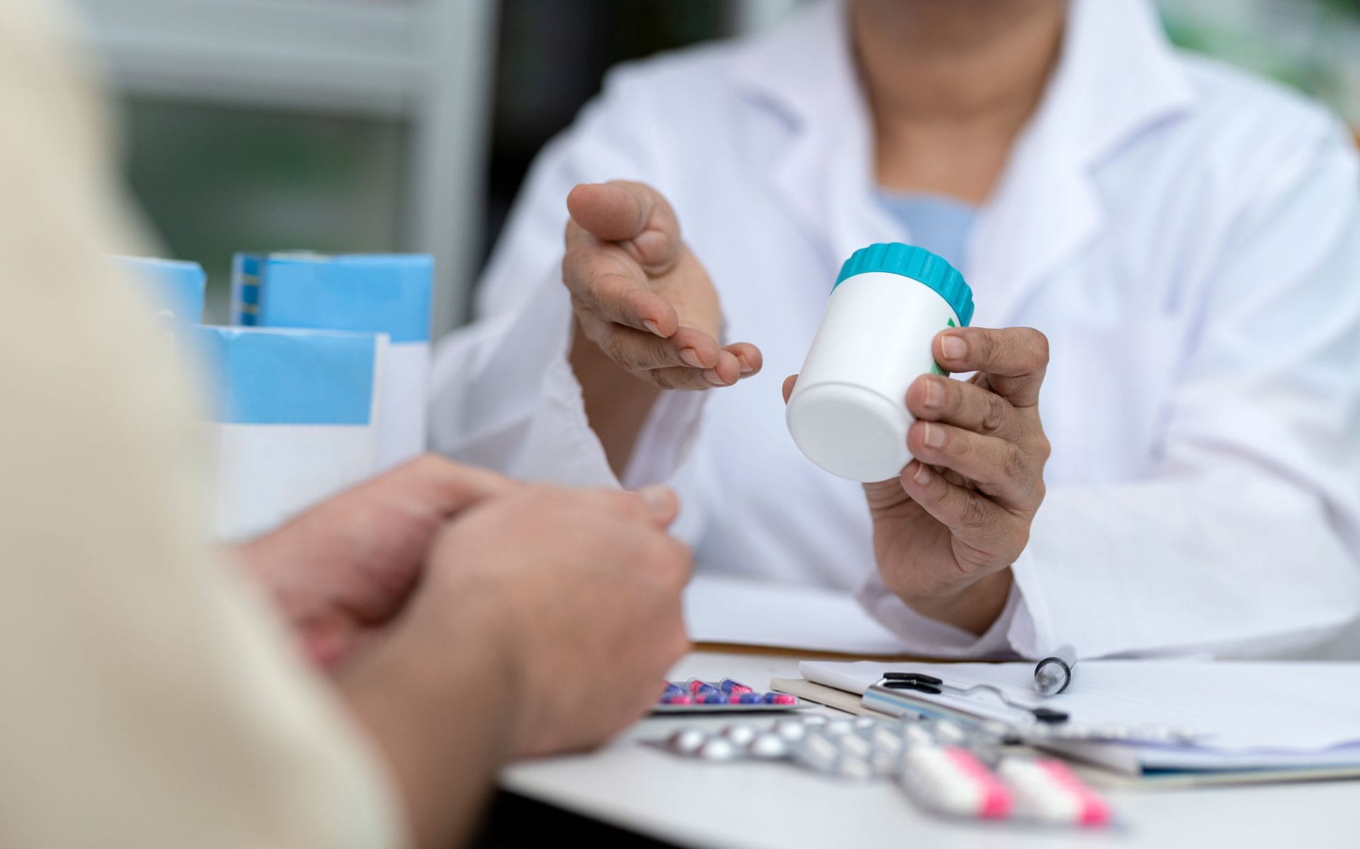 A pharmacist in a white coat offering a medication bottle to a patient, with a desk covered in various pill packs and a stethoscope in the foreground.