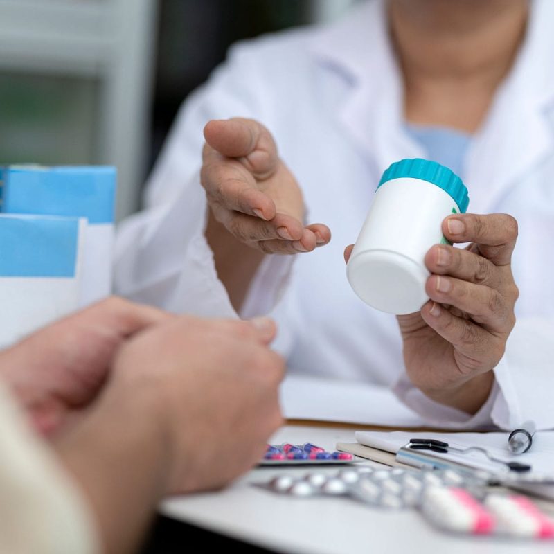A pharmacist in a white coat offering a medication bottle to a patient, with a desk covered in various pill packs and a stethoscope in the foreground.